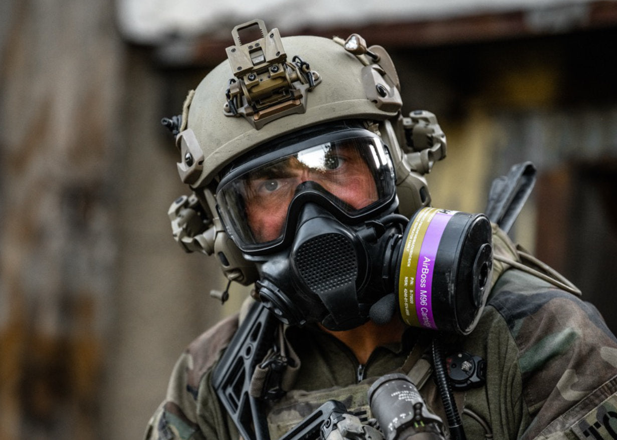 A close-up of a person wearing tactical gear, including a black gas mask, and a camouflage jacket, with a respirator canister.