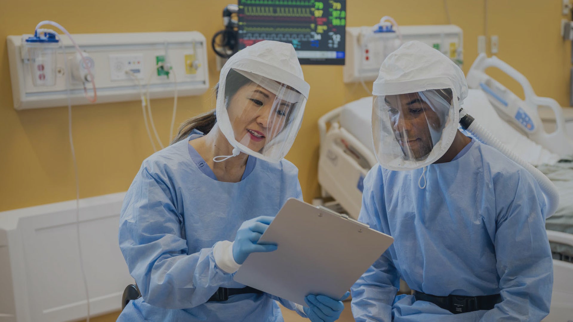 Two healthcare workers in protective suits and flexair face masks in a hospital room.